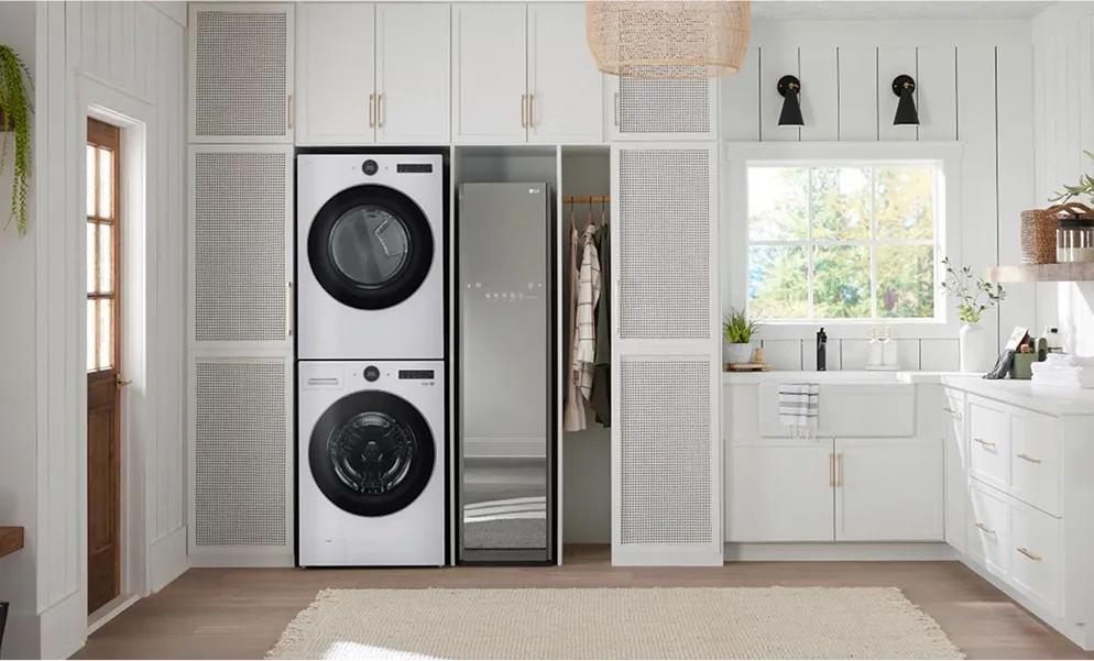 Stacked white front-load washer and dryer next to an LG Styler in a country-style laundry room with white cabinets and walls.