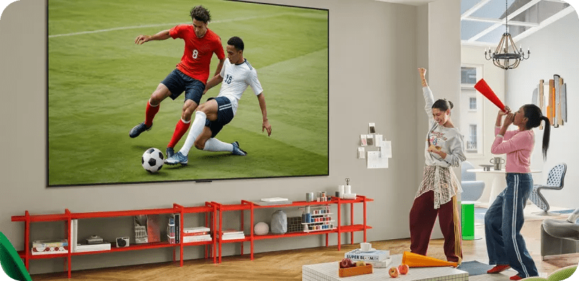 Two young women cheering a soccer match shown on a large wall-mounted LG TV in a bright living room with modern decor.
