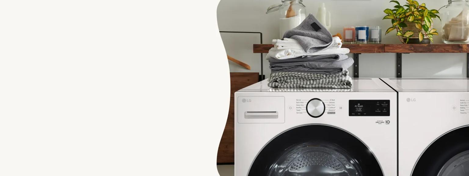 Close-up of a white LG front-load washing machine and matching dryer featuring a sleek control interface. A stack of folded grey and white laundry sits on the washer, with a rustic wooden shelf and plants in the background.