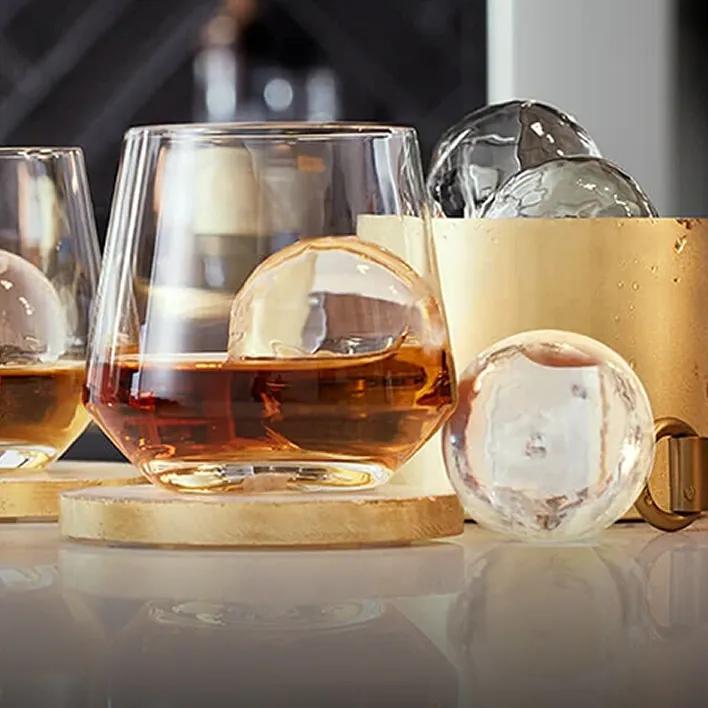 Close-up of a whiskey glasse partially filled with whiskey and containing a large spherical Craft Ice ball, with additional round ice spheres in a gold ice bucket and on the counter.