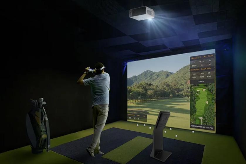 Person playing virtual golf in a simulation room with a projector screen displaying a golf course.