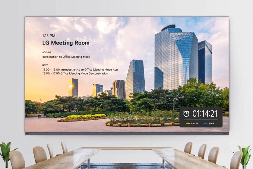 A corporate meeting space with a conference table and chairs. An LG 2K Ultimate Business Display is on the wall above the table, showing the day's meeting schedule over a bright image of a coastal city.