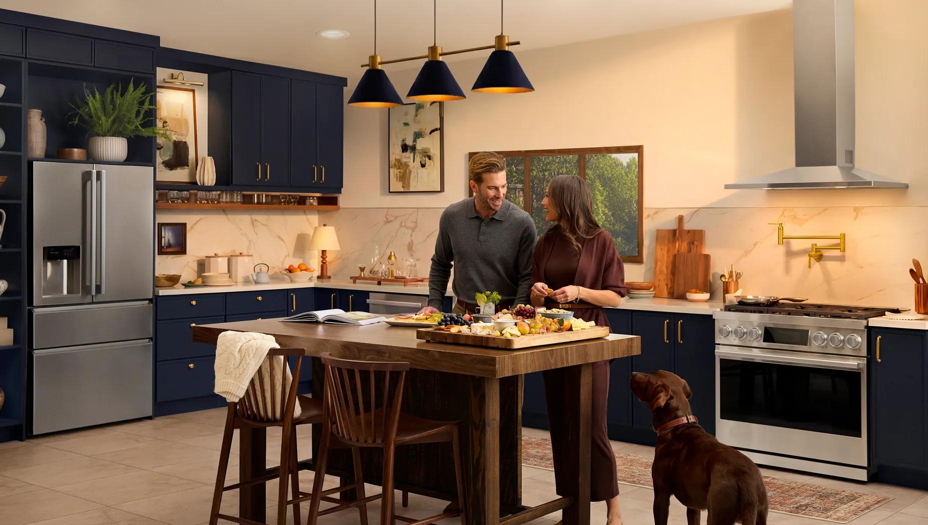 LG SIGNATURE Iconic Collection kitchen with refrigerator, range and wall-mounted range hood, in modern blue cabinetry, couple preparing food at island with dog nearby.
