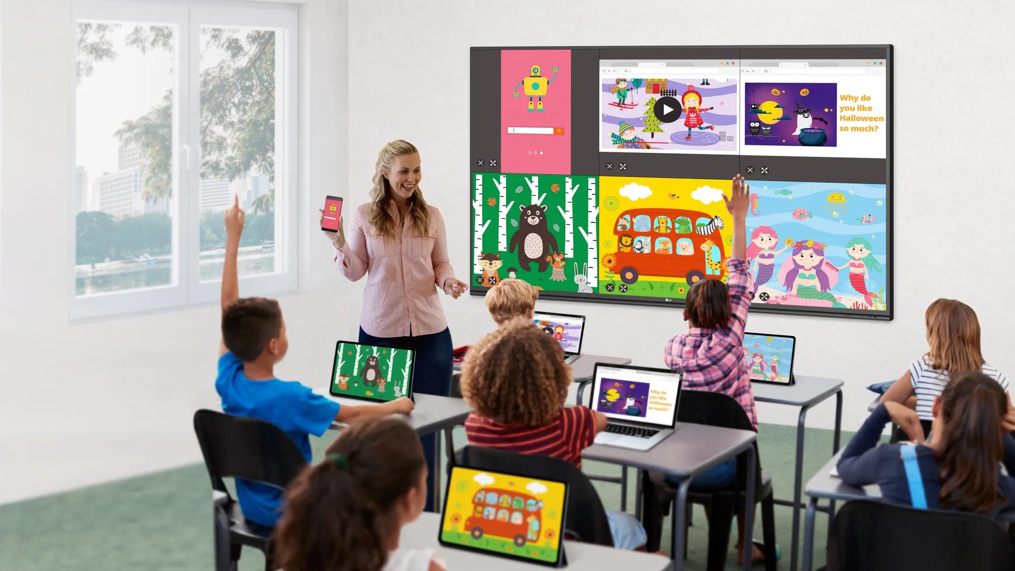 Teacher holding a smartphone leads a classroom lesson with an LG large-format display showing colorful educational content synced across student tablets and laptops.