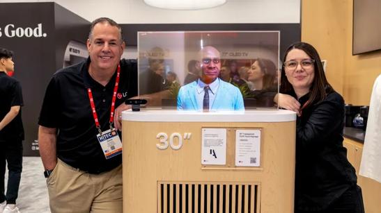 Two LG employees pose, smiling, on either side of an LG 30 inch transparent OLED digital display at HITEC 2025. The transparent display shows an image of a man in a professional shirt and tie.