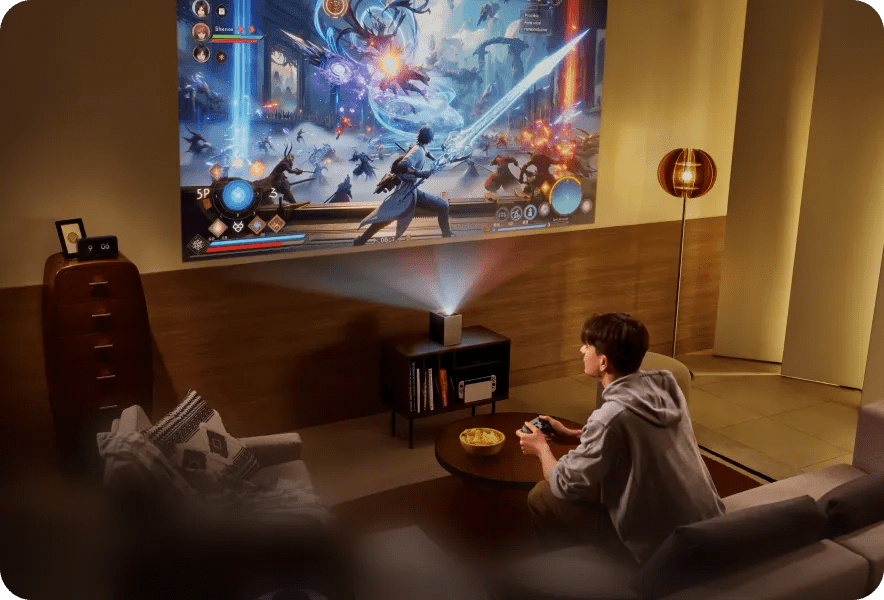 A young man sits on a sofa in a modern living room, holding a game controller while an LG CineBeam S projects a vivid fantasy battle scene on the wall.