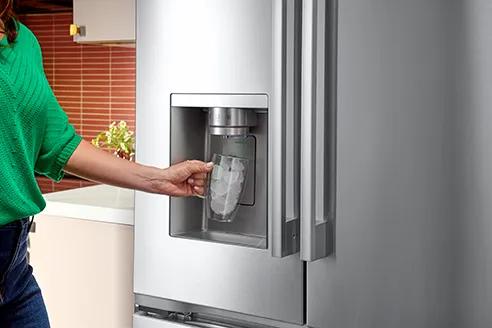 Woman with green sweater and blue jeans is filling a glass with ice from a French door refrigerator ice and water dispenser in a kitchen.