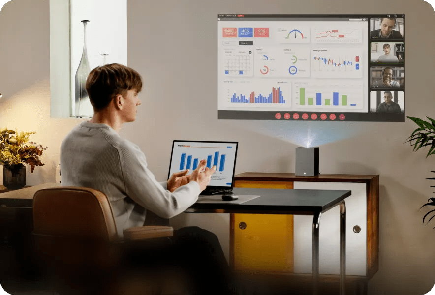 A young man works at a desk with a laptop while an LG CineBeam S displays charts, graphs, and a video conference call on the wall.