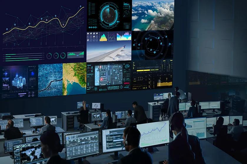 A control room with large screens displaying various data graphics and people working at computer stations.