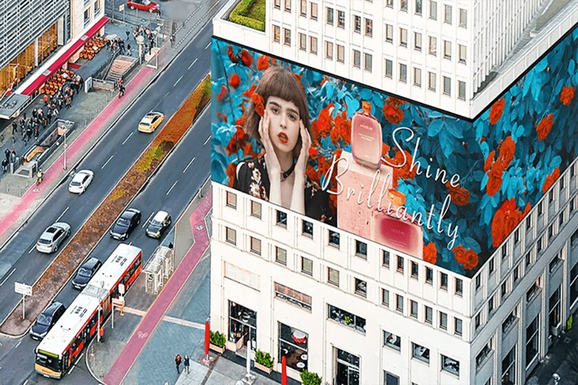 Aerial view of a street with a large building featuring a colorful advertisement of a woman and perfume bottles with the text "Shine Brilliantly."