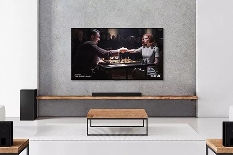 A set of 2 rear speakers subwoofer and a soundbar and TV are in a white living room A woman and a man are playing chess on TV screen