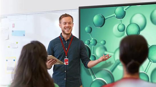 A man with a professional polo and lanyard gives a presentation to a small crowd. He's gesturing at a large LG display to his left.