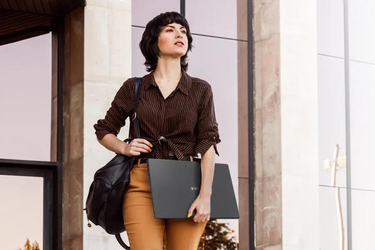 A young professional-looking woman standing in front of a city buliding. She's carrying a closed LG gram laptop in one hand. 