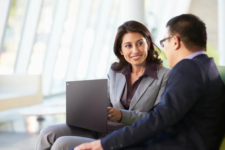 A businesswoman and businessman sit in a sleek office lobby, smiling as they chat. The woman has an LG ultraPC laptop, on her lap and is gesturing to something on the screen.