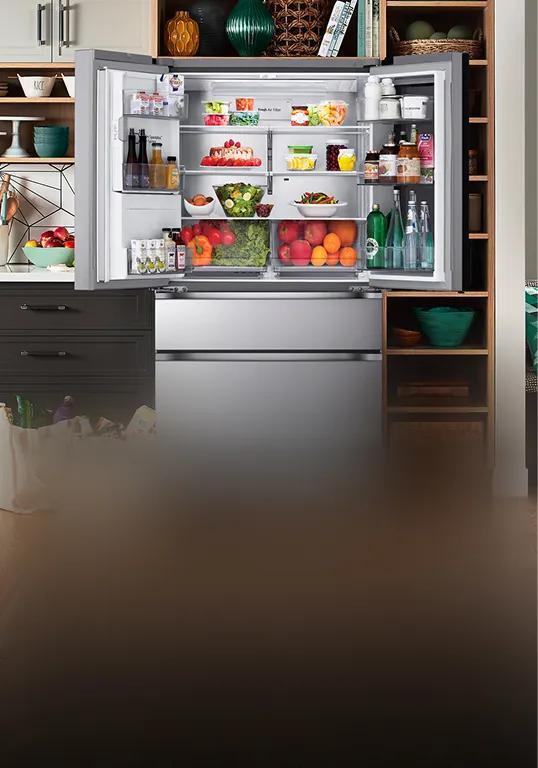 Open LG refrigerator in a modern kitchen fully stocked with produce, drinks, and groceries, with reusable shopping bags filled with food on the floor nearby.