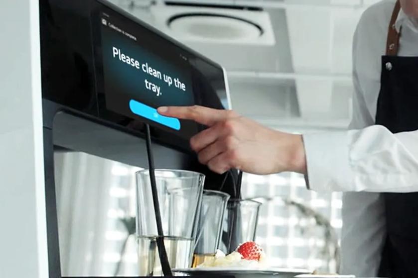 CLOi with dirty plates on her tray, with her display reading Please clean up the tray. A button reading OK is below that, which an employee is reaching out to press.