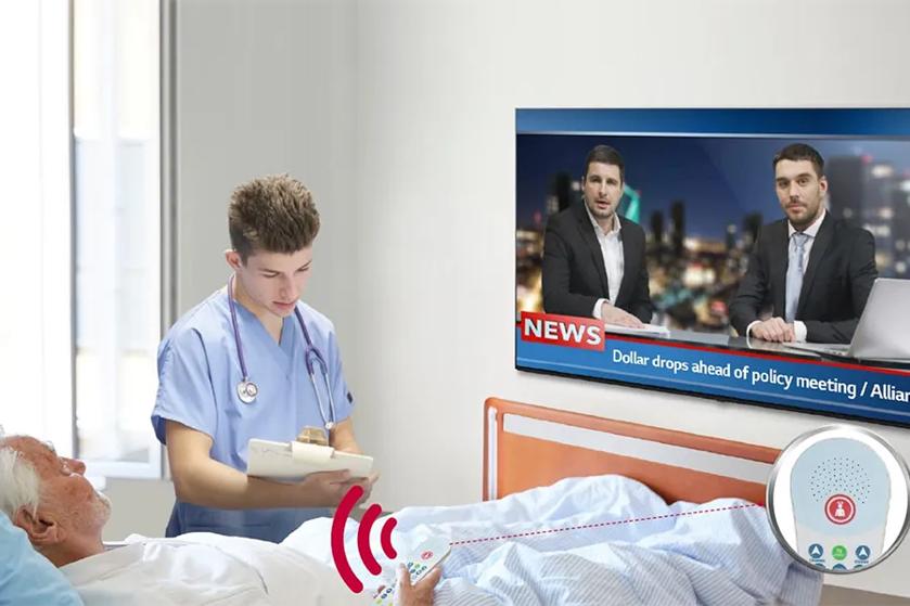 A nurse attends to a patient in a hospital room with a TV displaying a news broadcast. A nurse call button with a red light is active.