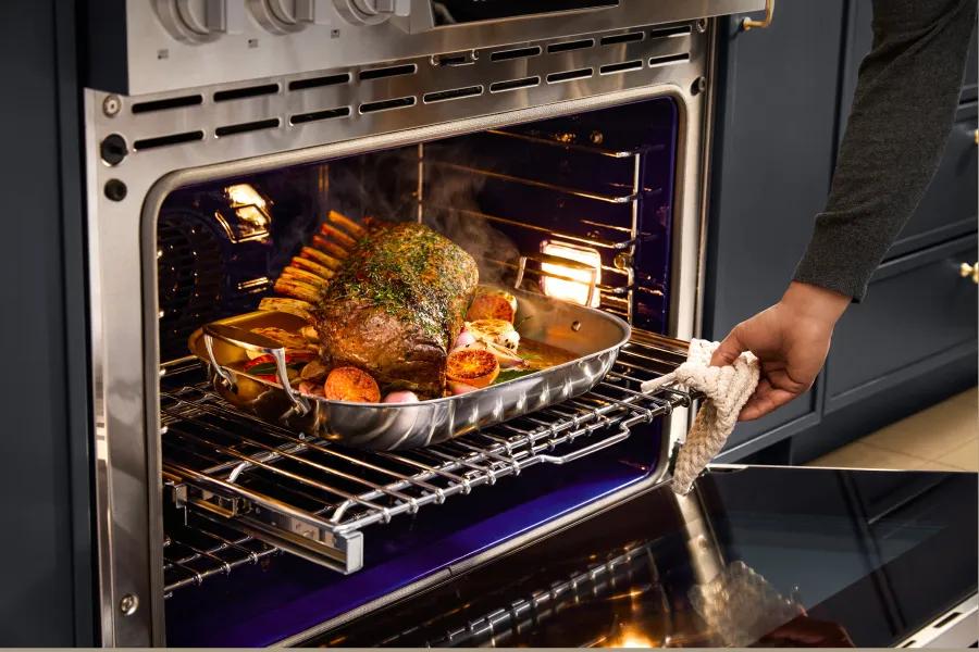 A person using a quilted pot holder to pull a sliding rack out of a modern LG oven, which contains a herb-crusted rack of lamb roasting in a stainless steel pan with citrus and vegetables.