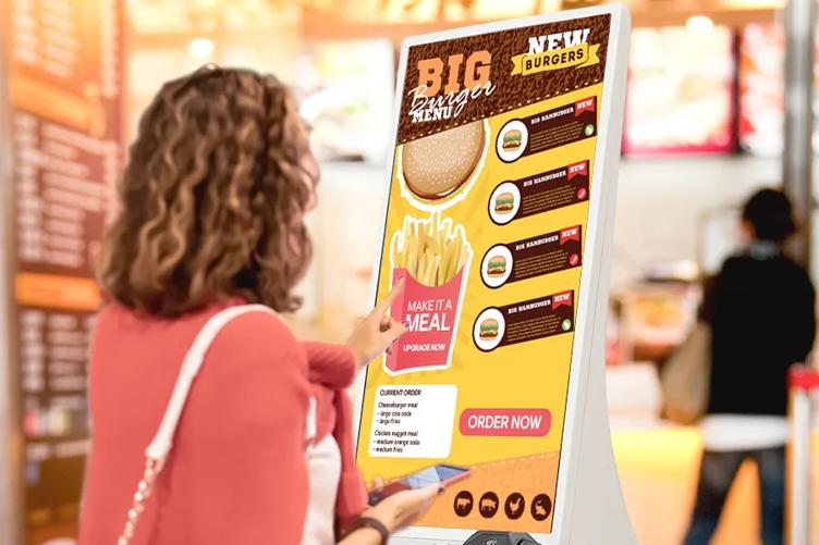 A woman stands in a brightly lit quick serve restaurant, using an LG Touch Kiosk to order a hamburger meal. 