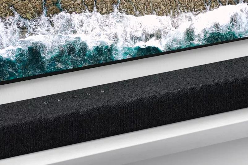 Close-up of a slight top view of LG Soundbar middle-to-right side Control buttons shown on middle of a product and LG logo shown on bottom right corner of a product Bottom side of TV is also visible waves at seaside are shown on TV screen Hi-Res AUDIO logo shown right below the product