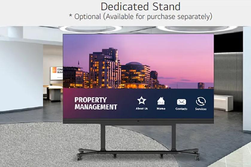 The image shows a large digital display on a stand in a modern indoor setting. The screen features a cityscape at dusk with buildings lit against a pink and purple sky. The lower section of the screen displays the text "PROPERTY MANAGEMENT" on the left and icons with labels for "About Us," "Home," "Contacts," and "Services" on the right. The display is mounted on a metal stand with wheels.