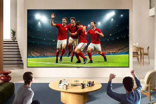 LG TV for sports fans is wall-mounted in a bright living room, showing a soccer celebration with vivid colors and refined picture quality across a wide screen, as a family seated on sofas cheers together.