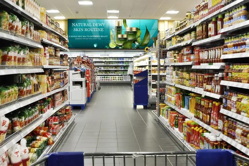 A shopping cart being pushed down a store aisle. At the end of the aisle, a large video wall advertises beauty products.