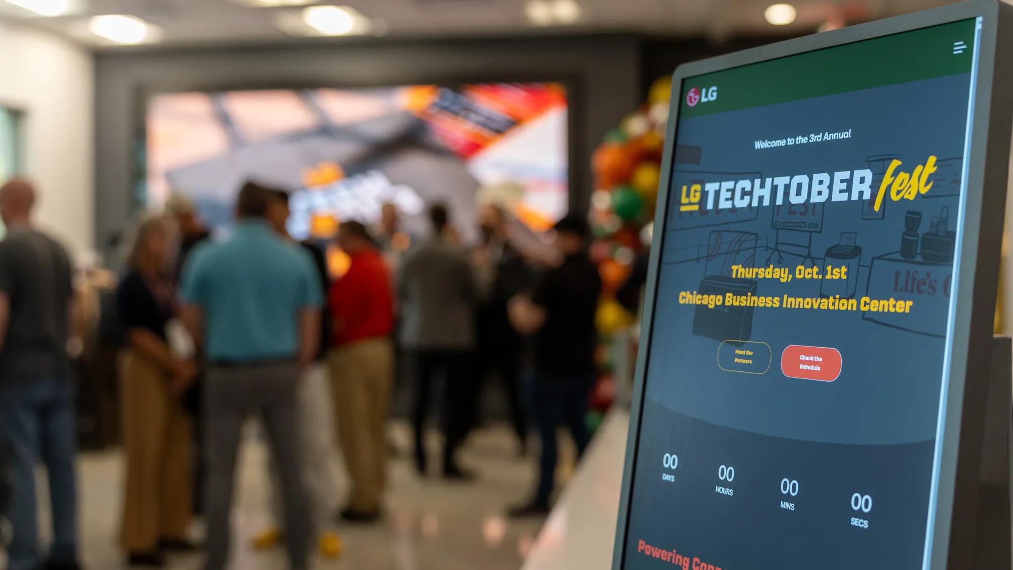 A vertical LG kiosk screen displaying the 'Welcome to the 3rd Annual LG Techtober Fest' page, Thursday, Oct. 1st at the Chicago Business Innovation Center, with a countdown timer and 'Meet Our Partners' and 'Check the Schedule' buttons. Attendees mingle in the blurred background.