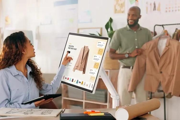 Two people in an office, one seated pointing at a computer screen displaying a jacket, the other standing with a similar jacket.