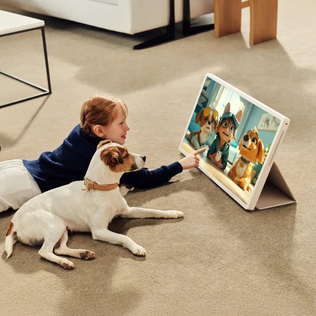 A young girl lying on a carpet smiling and pointing next to a dog, both watching an animated movie on a large StandbyME 2 screen propped up on a stand. The screen shows three animated characters—two dogs and a child