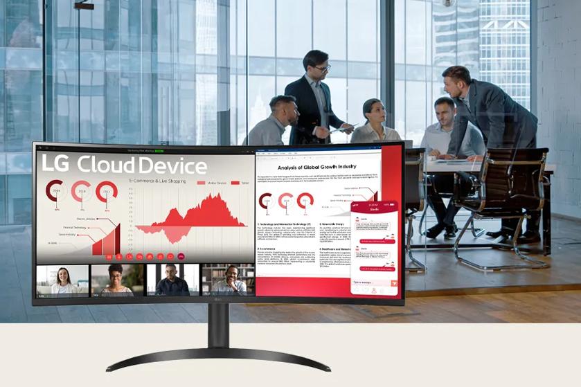 Office scene with a curved monitor showing graphs, text documents, and video call participants, with people meeting in the background.