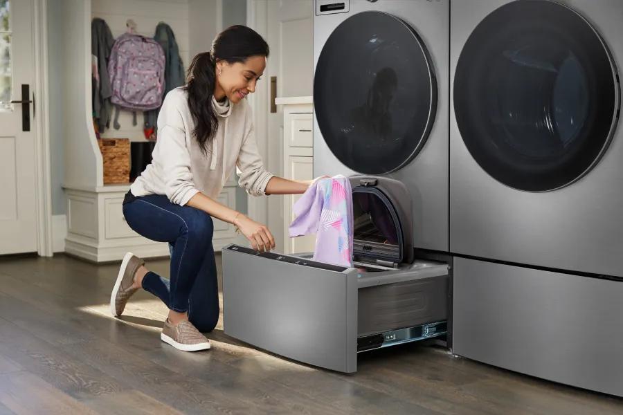 A woman loads clothes into an LG Sidekick pedestal washer beneath a front-load washer and dryer in a bright laundry room.