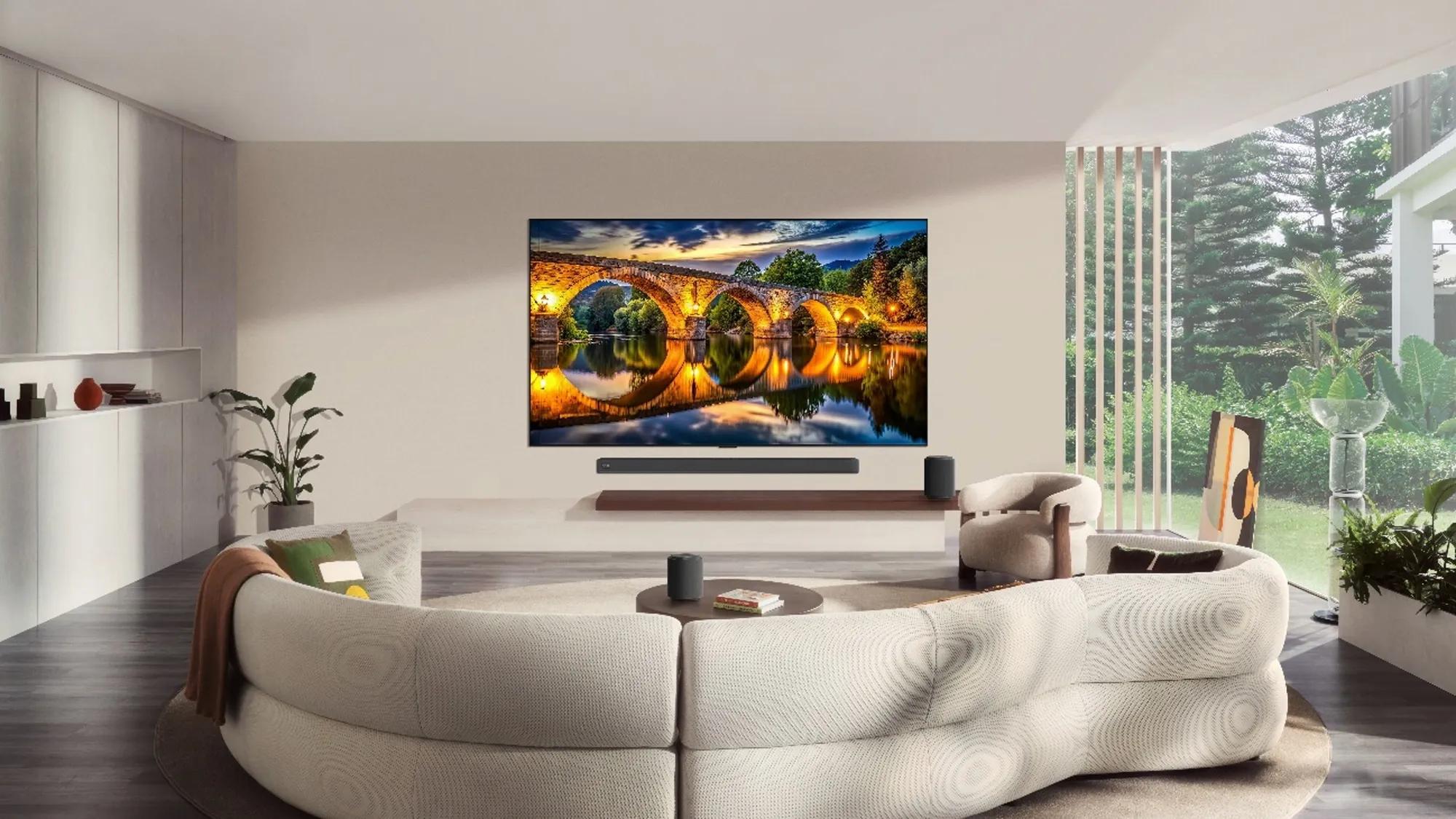 A wall-mounted TV shows a glowing stone arch bridge reflected in a river at dusk, above a soundbar, with wireless speakers around a curved white sofa.