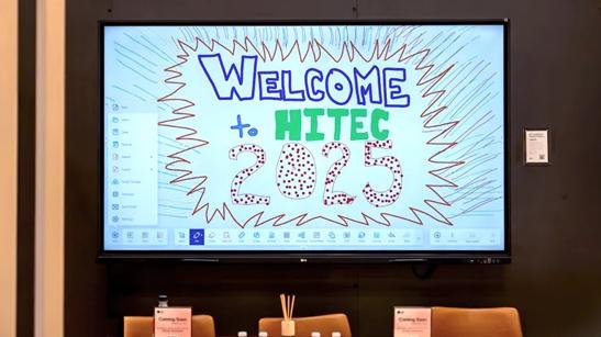 An LG CreateBoard smart board display is mounted in the LG HITEC booth above a meeting table. The screen displays a message written using the touch screen capabilities, in various colors and patterns. The message reads Welcome to HITEC 2025.
