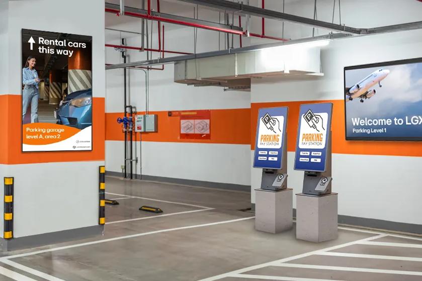 A parking garage with LG self ordering kiosks displaying ticket payment options on their screens, and signage welcoming visitors to the airport and indicating the location of rental vehicles.