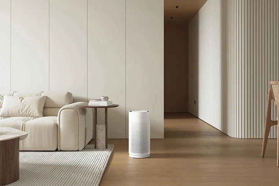 Slim white cylindrical air purifier positioned on wooden floor in minimalist living room with cream sofa and neutral decor