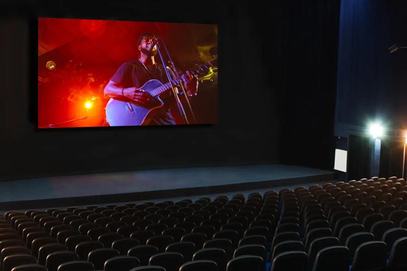 Movie theater showing a performer playing guitar and singing on screen, with rows of empty seats.