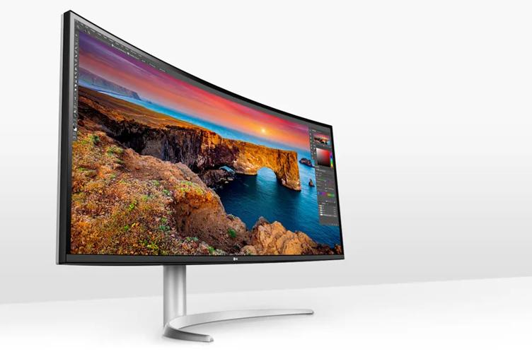 An LG Ultra Wide Curved monitor on a smooth white desk. An editing software is open on its display, visible in the top ribbon, a panel for a color selector, and a layers panel, but most of the monitor's display is taken up in a bright nature photograph of sea cliffs at sunset.