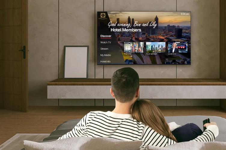 A couple in a hotel room relax in bed while watching an LG Hotel TV. The TV is open to a menu greeting them by name, with options along the left of the screen that read: Discover, Watch TV, Stream, My Media, and Hotel Info. Discover is bolded and underlined to indicate that it's currently selected. Three small picture-in-picture screens are below the welcome message, with captions reading: Your hotel, Travel spotlight, and Local news.