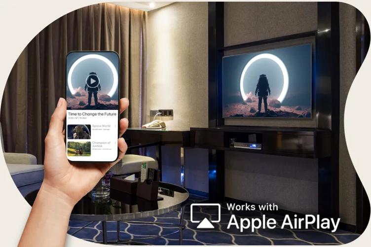 An LG Hotel TV is in an upscale hotel room. A movie with an astronaut is displayed on the screen. A hand holds a smartphone in the forefront of the scene, with the same astronaut movie on its display. Text in the corner of the image reads Works with Apple Airplay with an airplay icon.
