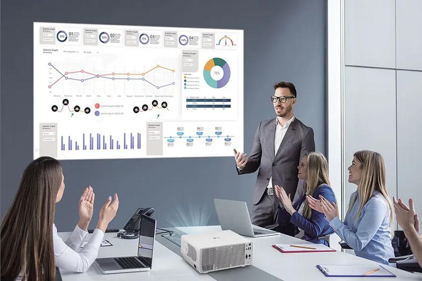 The image depicts a modern office meeting room where a group of four people is engaged in a presentation. In the center, a man in a suit is standing and gesturing towards a large projection screen displaying various charts and graphs. Three women are seated at a conference table, clapping and looking towards the presenter, suggesting that they are engaged and appreciative of the presentation. The background features a wall with a large screen displaying colorful line graphs, pie charts, and bar graphs, as well as numeric statistics. A projector is placed on the table, projecting the image onto the screen. Additional items like notebooks and pens are present, indicating an environment set for a business meeting.