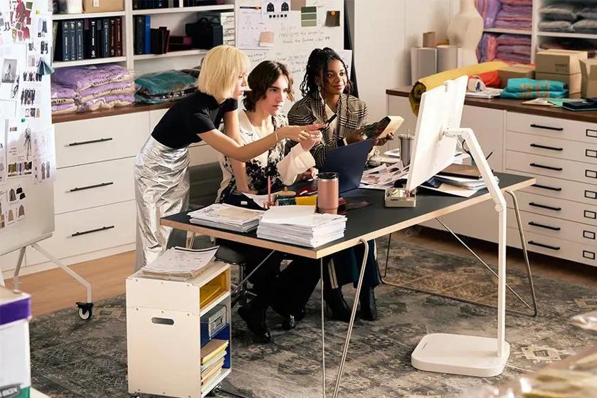Three people collaborate around a table covered with papers and a monitor, in a studio with textiles and sketches in the background.