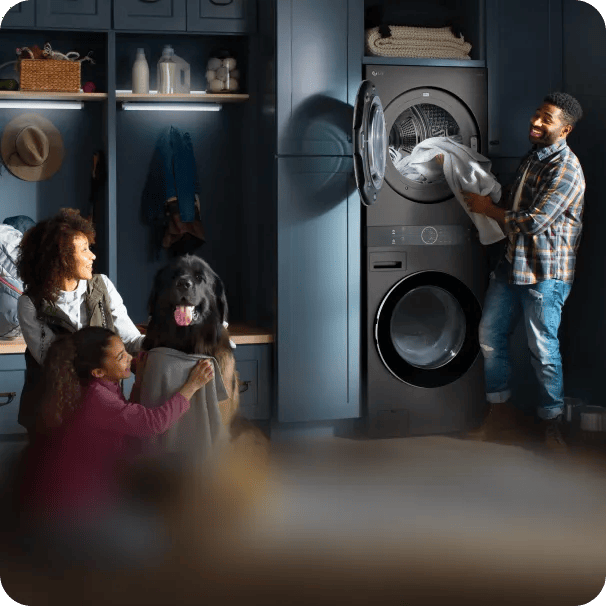 A smiling man standing in a laundry area, pulling clean white linens from the top drum of a dark metallic LG WashTower stacked washer-dryer, while a black dog watches from the side.