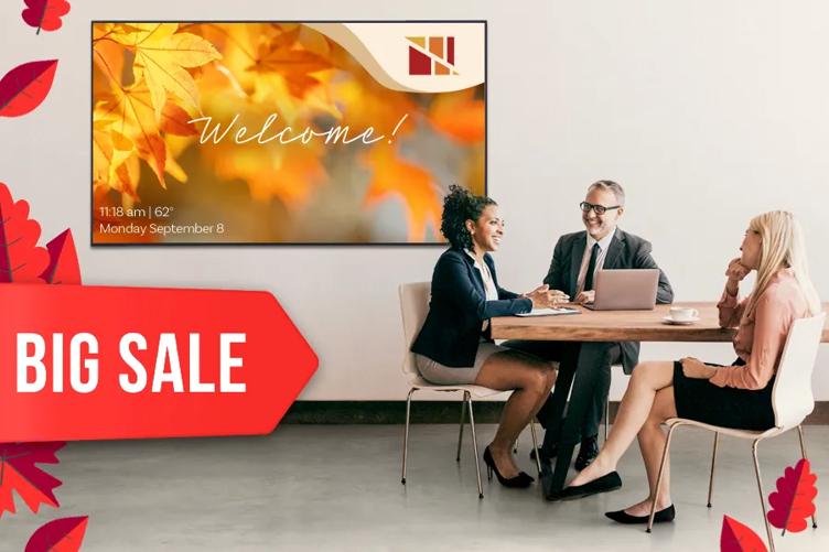Three business professionals sit at a small table in an office lobby, smiling as they chat. On the wall behind them, an LG Digital Signage displays a fall graphic welcoming guests to the office. A large red tag in the bottom left corner of the image reads Big Sale and is surrounded by red cartoon fall leaves.