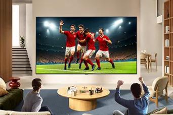 LG TV for sports fans is wall-mounted in a bright living room, showing a soccer celebration with vivid colors and refined picture quality across a wide screen, as a family seated on sofas cheers together.