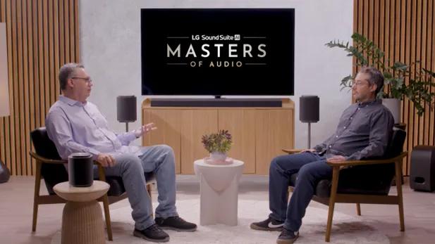 Two men talk in armchairs on either side of a TV that reads “LG Sound Suite AI Masters of Audio,” with a soundbar and surround speakers around the set.
