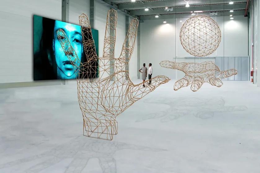 A spacious room with wireframe sculptures of hands and a sphere, and a digital screen showing a blue-toned face.