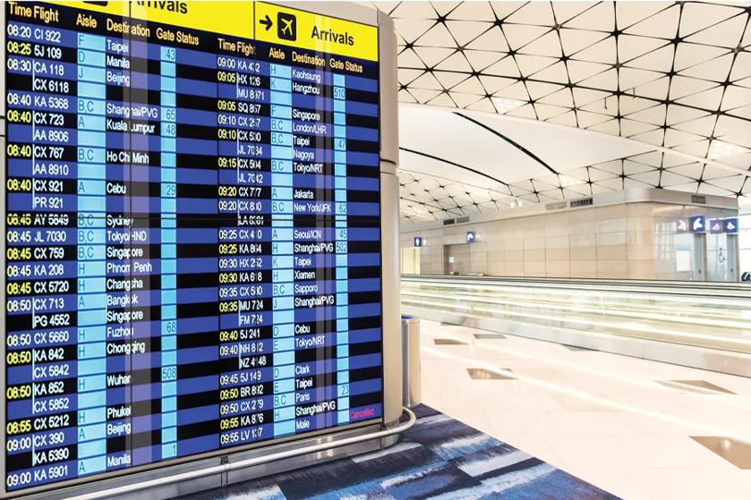 A large signage display in an airport terminal shows the latest arrival and departure times.