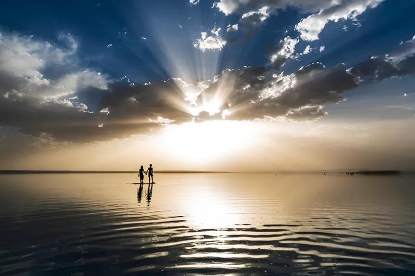 Two people stand in shallow water beneath a cloud-filled sky as the low sun casts rays and reflects across the rippling surface.