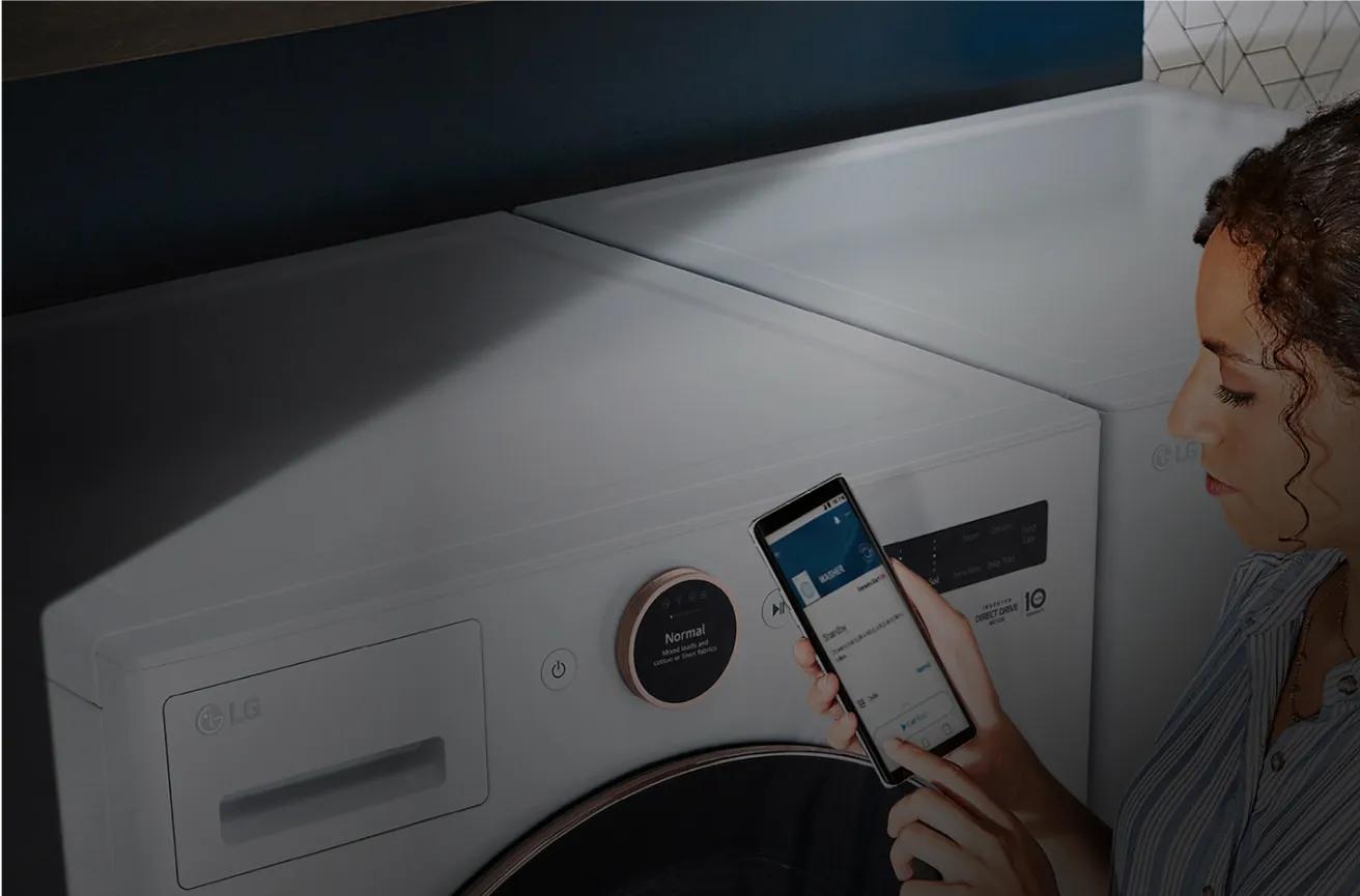 Woman standing in front of her washer and try with the ThinQ app on her phone and LG ThinQ logo at the top right.